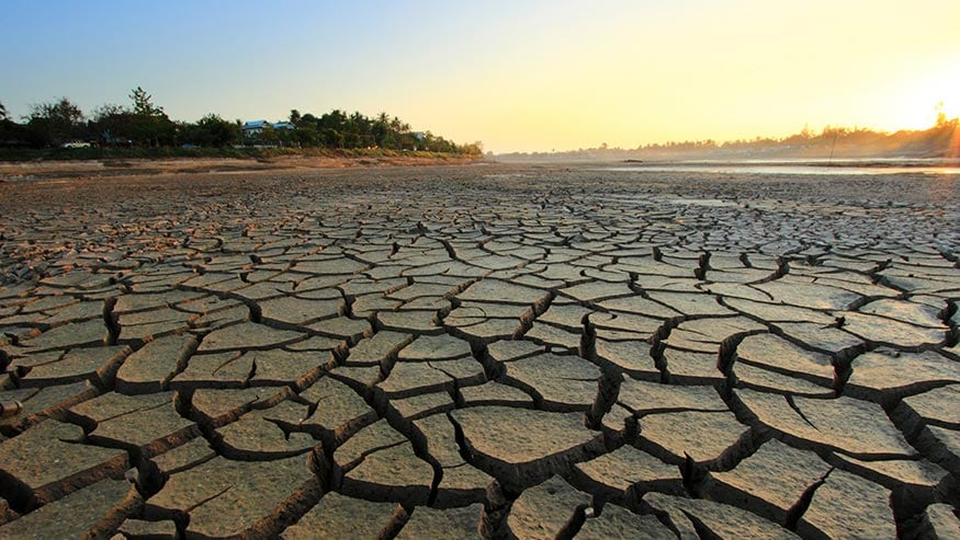 Outdoor landscape with the sun on the horizon, and a cracked and parched earth in the forefront.
