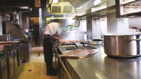 Chef bent over a broiling station working on cleaning and sanitizing equipment in commercial kitchen.