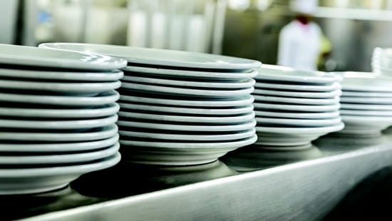 Five stacks of newly cleaned porcelain dinner plates sitting on a steel ledge in a commercial kitchen.